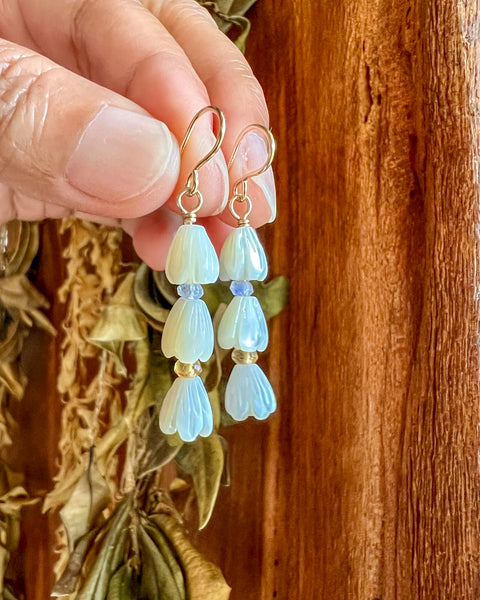 Stack of 3 carved mother of pearl pikake bud with moonstone & citrine rondel earrings held by a fingertips against a pig board with dried lei background