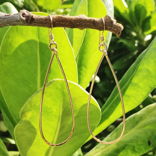 These hoop earrings feature an eye-catching slender teardrop shape with a flattened, light-catching hammered finish along the bottom edge. Metal: 14K Gold-Filled Length: 2.5" aprox. Earrings displayed in front of naupaka bush.